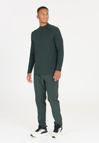 Donkergroene langetermijn top met een halve rits kraag, gecombineerd met bijpassende donkergroene broek en zwarte sportschoenen met witte accenten.