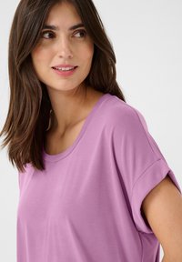 Gaiši violets īsām piedurknēm t-krekls ar aplīgu kakla izgriezumu un sarullētām piedurknēm, izgatavots no gludas auduma, kas mīksti krīt.