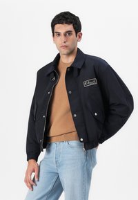 Marineblauer Bomberjacke mit Reißverschluss vorne, gerippten Bündchen und Seitentaschen. Mit einem kleinen gestickten Markenlogo auf der Brust.