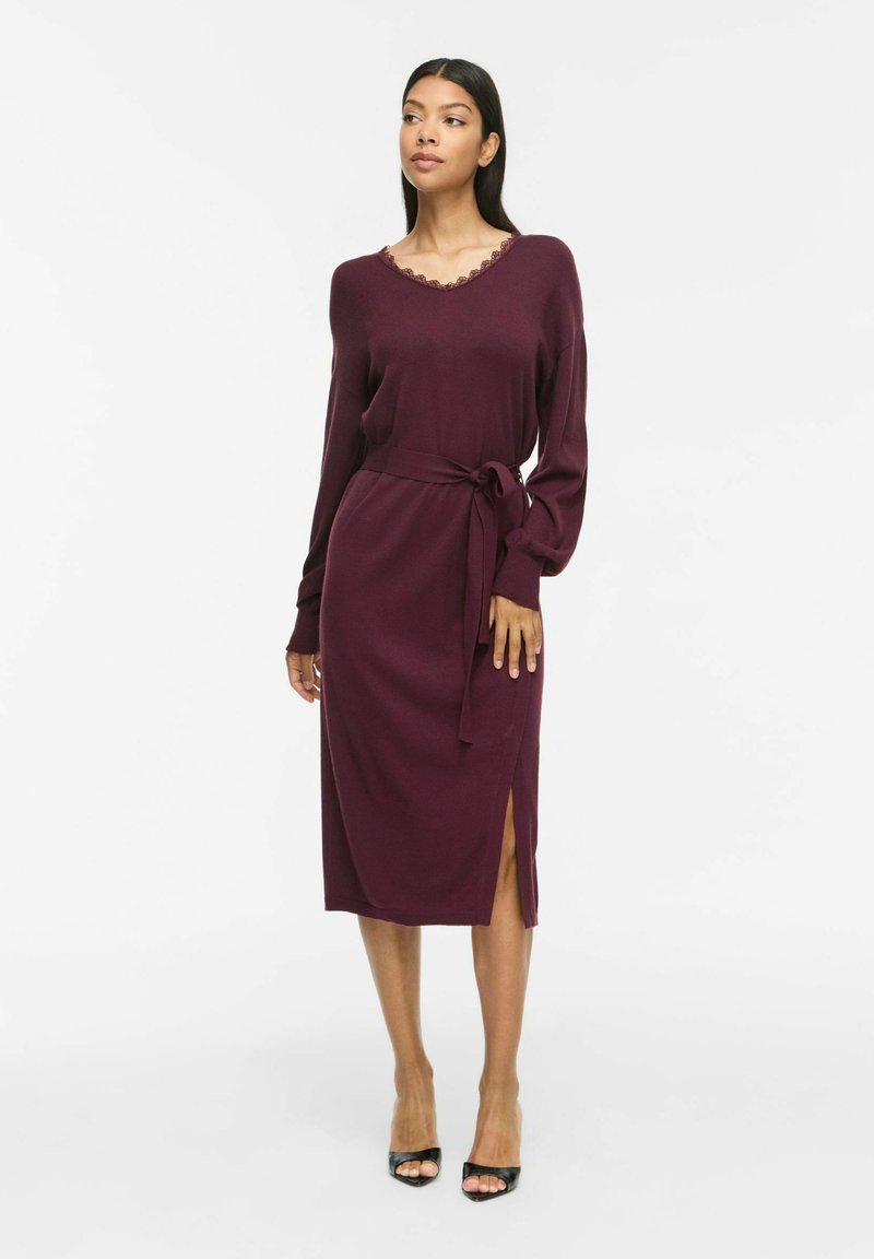 Femme portant une robe midi bordeaux à manches longues avec une ceinture à la taille et une fente latérale, associée à des talons ouverts noirs, debout sur un fond blanc.