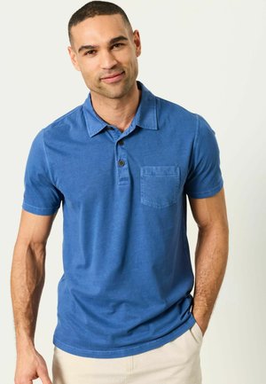 Man with short hair wearing a blue short-sleeve collared shirt with buttons and a chest pocket, standing with one hand in beige pants pocket.