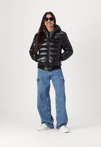 Zwarte gewatteerde jas met capuchon, glanzende textuur, voorrits, zwarte tailleband; gecombineerd met lichtblauwe wijde jeans en witte sneakers.