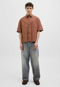 Camisa de cuadros de manga corta en óxido y beige, con cierre frontal de botones y un bolsillo en el pecho, combinada con jeans de gran tamaño en azul claro.