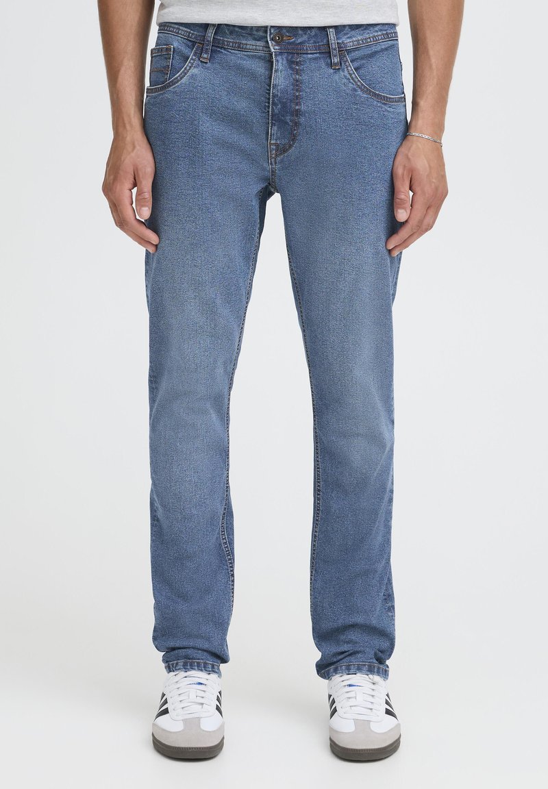 Zilas džinsa bikses ar šauru piegriezumu, pieciem kabatām, vieglu nodiluma efektu un kontrastējošām šuvēm, kopā ar baltām sporta kurpēm.