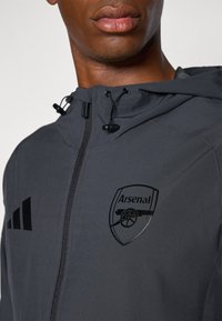 Grå hettejakke med svarte detaljer, har et preget Arsenal-logo og glidelåslukking, laget av et glatt, lett materiale.