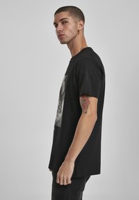 Sort bomulds T-shirt med et stort grafisk tryk på forsiden. Korte ærmer, løs pasform og flad kant. Huden tonede tatoveringer synlige på armen.