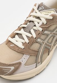 Beige og brun løpesko med mesh- og mockaskinnoverdel, kremfargede lisser og grå detaljer. Har en polstret såle og et særegent logo.