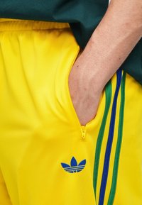 Żółte spodenki sportowe z kieszenią na zamek, zdobione niebiesko-zielonymi paskami oraz niebieskim logo Adidas na udzie. Gładka faktura.