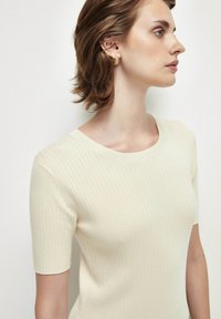 Top de manga corta con canalé en suave tejido color crema, con un escote redondo y una textura sutil. Modelo con perfil lateral.