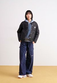 Schwarze gesteppte Jacke mit Druckknöpfen über einem hellblauen Hoodie, kombiniert mit lockeren marineblauen Cargo-Hosen und weißen Sneakers. Einfaches, funktionales Design.