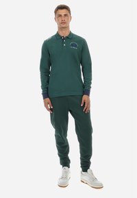 Zelena dolga polo srajca s ovratnikom, z zapenjanjem na tri gumbe in detajlom logotipa. Združena z ujemajočimi se trenerkami. Bele športne copate za popoln videz.