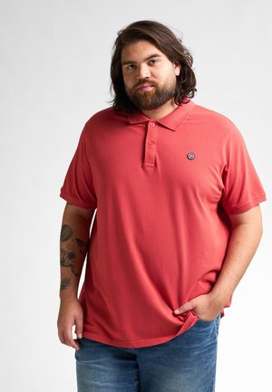 Polo shirt corail avec un col et une patte de boutonnage à trois boutons. Il dispose de manches courtes et d'un petit logo sur la poitrine gauche. Jean en denim.