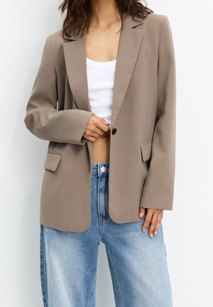 Kvinde iført beige blazer over hvid crop top og lyseblå højtaljede jeans mod en ensfarvet baggrund.