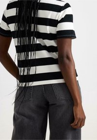 Camisa de rayas en blanco y negro de manga corta con dobladillo redondeado, combinada con pantalones de talle alto en gris oscuro. Se ve el cabello trenzado.