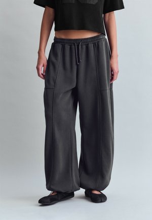 Personen bærer løstsiddende mørkegrå joggingbukser med elastisk talje og snøre, kombineret med sorte split-toe sko og en cropped sort top.