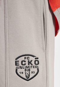 Grå hettegenser med en rød erme, med en brodert svart logo, "FC ECKŌ UNLIMITED," og et grafisk bilde av et hode inne i et skjold.