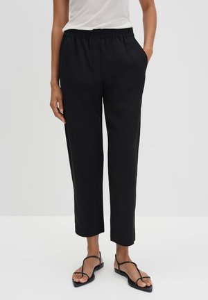 Kobieta w czarnych, krótkich spodniach z elastyczną talią i czarnych, płaskich sandałach na paski, stojąca na jednolitym, jasnym tle.