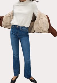 Suéter de punto beige claro, jeans rectos azules, abrigo forrado de borrego marrón y bailarinas negras con punta redonda.