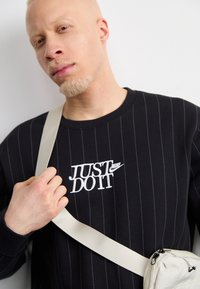 Fekete csíkos pulóver fehér "JUST DO IT" Nike logóval, bézs válltáskával, amely cipzáros záródású és pánttal rendelkezik.