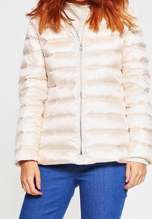 Nainen käyttää kiiltävää beigea toppatakkia valkoisen neulotun villapaidan ja sinisten farkkujen päällä, seisoo rentoina käsivarsina sivuilla.