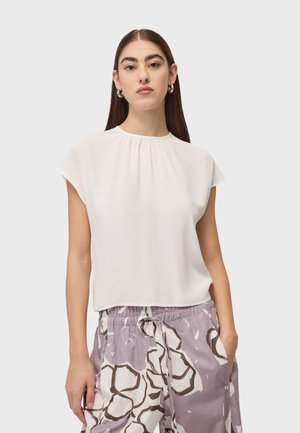 Femme portant un haut blanc sans manches et un pantalon à cordon de serrage violet clair avec des motifs floraux foncés, debout avec une main dans la poche.