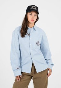 Blau-weiß gestreiftes Langarmhemd mit Knopfleiste, Brusttasche und besticktem Detail. Getragen mit braunen Hosen und einer schwarzen Mütze.