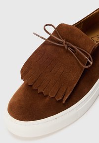 Mocassins en daim marron avec une semelle en caoutchouc blanche, un rabat frangé décoratif et un nœud en cuir attaché sur le dessus.
