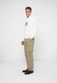 Felpa bianca con logo "H" nero, abbinata a pantaloni cargo beige con molteplici tasche e scarpe nere. Design semplice e funzionale.