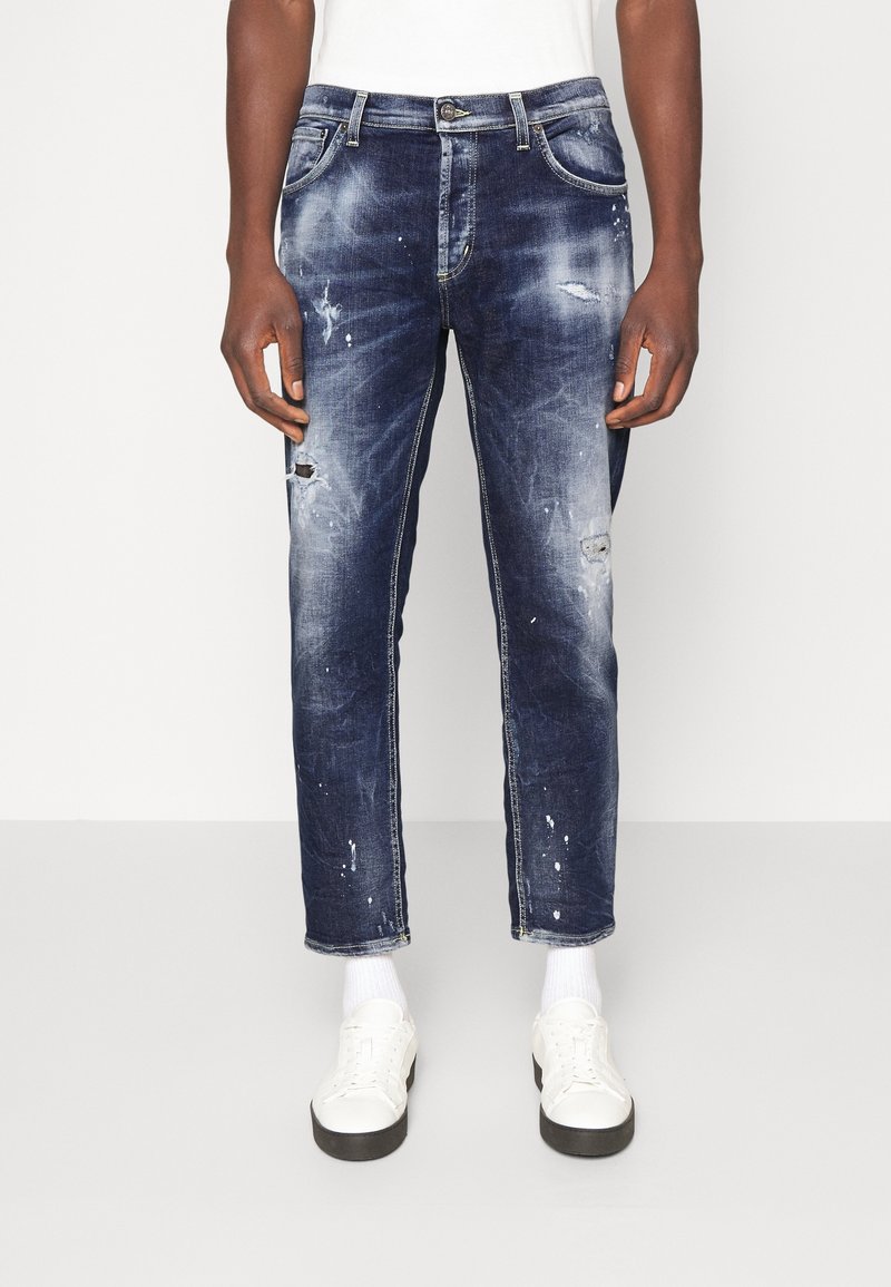Homme portant un jean bleu foncé usé avec des éclaboussures de peinture, des chaussettes blanches et des baskets blanches, debout devant un fond uni.