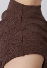 Κοντινό πλάνο στον ώμο και τον λαιμό με ένδυμα καφέ χρώματος, υφασμένο με ανάγλυφη υφή, ψηλό λαιμό και ορατές κάθετες ραφές.