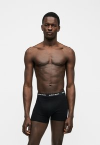 Μυώδης άνδρας σκουρόχρωμος, στέκεται με το πρόσωπο προς τα εμπρός, φορώντας μαύρα μποξέρ Björn Borg σε απλό ανοιχτό φόντο.