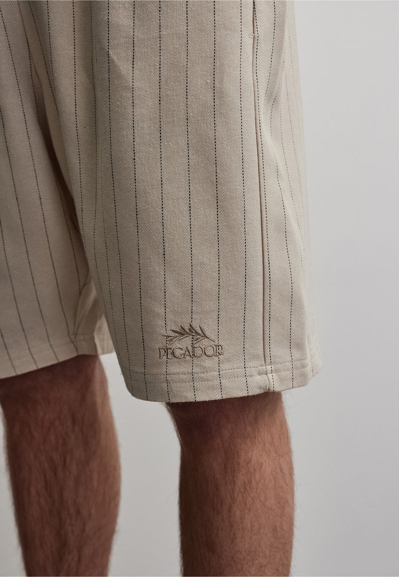 Short beige à fines rayures verticales porté par une personne, avec le logo « PEGADOR » brodé près de l'ourlet sur la jambe gauche.