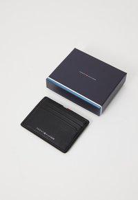 Black leather cardholder with textured surface, featuring multiple card slots and embossed logo, accompanied by a navy box with a striped detail.