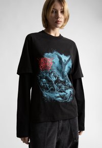 Melna īsām piedurknēm T-krekls ar zilu grafisko mākslu, kurā attēlota eņģelis un figūras. Augšā ir sarkanas krāsas teksta detaļa. Uzvilkts virs garām piedurknēm krekla.