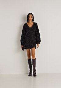 Melns ziedu raksts kleitā ar garām balona piedurknēm un V-veida kakla izgriezumu, apvienota ar augstiem brūnu ādas zābakiem. Tur rokās mazu melnu somiņu.