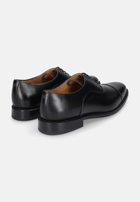 Ein Paar schwarze Leder-Herrenschuhe mit Schnürung und Kappenzehen-Details, seitlich von hinten auf einem weißen Hintergrund gezeigt.
