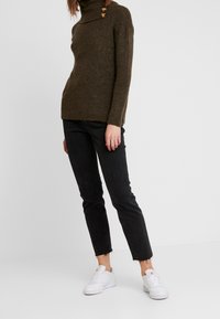 Pull en maille vert olive avec col roulé et trois boutons décoratifs, associé à un jeans noir coupé et des baskets blanches avec un logo rouge.