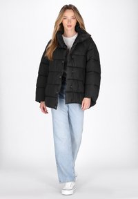 Kvinde iført en sort puffersjakke, hvid skjorte, lyseblå jeans og hvide platfromsko går mod kameraet foran en hvid baggrund.