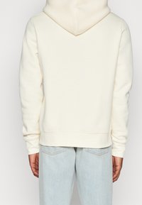 Felpa color crema con cappuccio regolabile, polsini e bordo a costine. Il tessuto sembra morbido e liscio, abbinato a jeans azzurri.