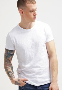 Hvid bomuld t-shirt med korte ærmer, rund hals, og en let tekstur. Har farverige tatoveringer på modellens venstre arm.