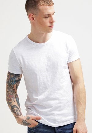 White cotton t-shirt with short sleeves, crew neckline, and a slight texture. Features colorful tattoos on the model's left arm.