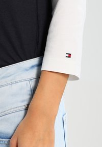 White long-sleeve shirt with a navy blue torso and a small embroidered Tommy Hilfiger logo. Paired with light blue denim jeans.