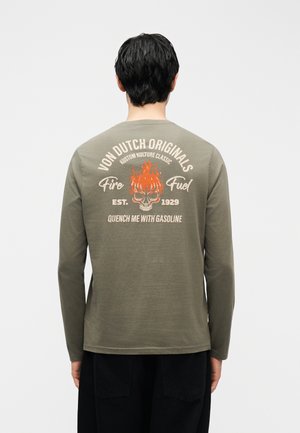 Mees, kes kannab oliivrohelise pika varrukaga särki tuleleviga kolbajäga kujutise ja tekstiga "Von Dutch Originals Fire Fuel Est. 1929" seljal, selg pööratud vaatajast eemale.