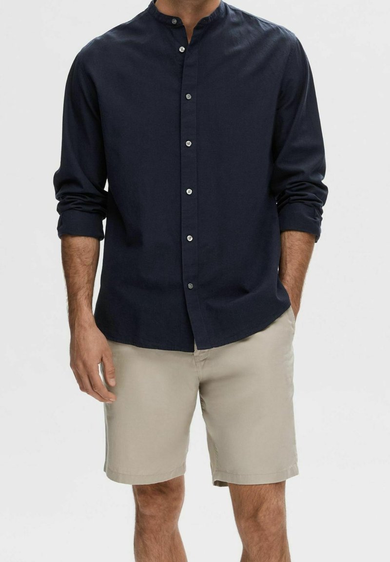 Chemise bleu marine à col montant, à manches longues et boutons argentés, associée à un short beige en tissu léger.