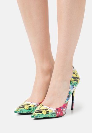 Jambes portant des chaussures à talons hauts colorées avec des motifs de graffiti abstraits en jaune, vert, rouge et bleu sur un fond blanc.