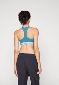 Ciklast modra športna srajca z zasnovo racerback, ki vsebuje mehak elastični pas z oznako "new balance." Uporabljena z črnimi širokimi hlačami.