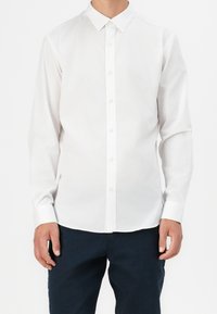 Homme portant une chemise blanche unie à manches longues avec col et un pantalon foncé sur fond blanc.
