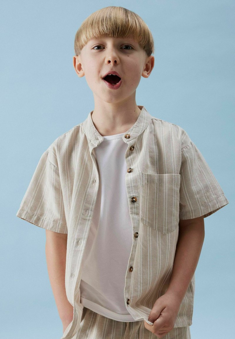 Camicia a manica corta a righe beige e bianca con colletto, dotata di bottoni in legno e una tasca frontale, abbinata a una t-shirt bianca sotto.