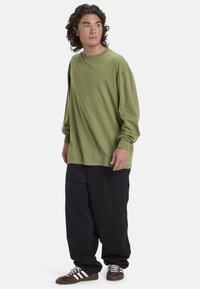 Langarm-Olivgrün T-Shirt mit Rundhalsausschnitt, lockere Passform, kombiniert mit lockeren schwarzen Hosen und braunen Schuhen mit weißen Streifen.