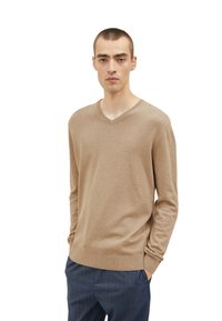 Beige V-Ausschnitt-Pullover aus weichem Stoff, mit langen Ärmeln und gerippten Bündchen. Kombiniert mit dunklen, maßgeschneiderten Hosen. Minimalistisches Design.